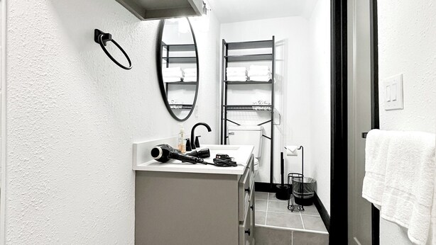A compact bathroom with a built-in sink, a mirror, a towel rack, and a shelf with folded towels. - Houston Home by NRG Stadium & Downtown (Houston)