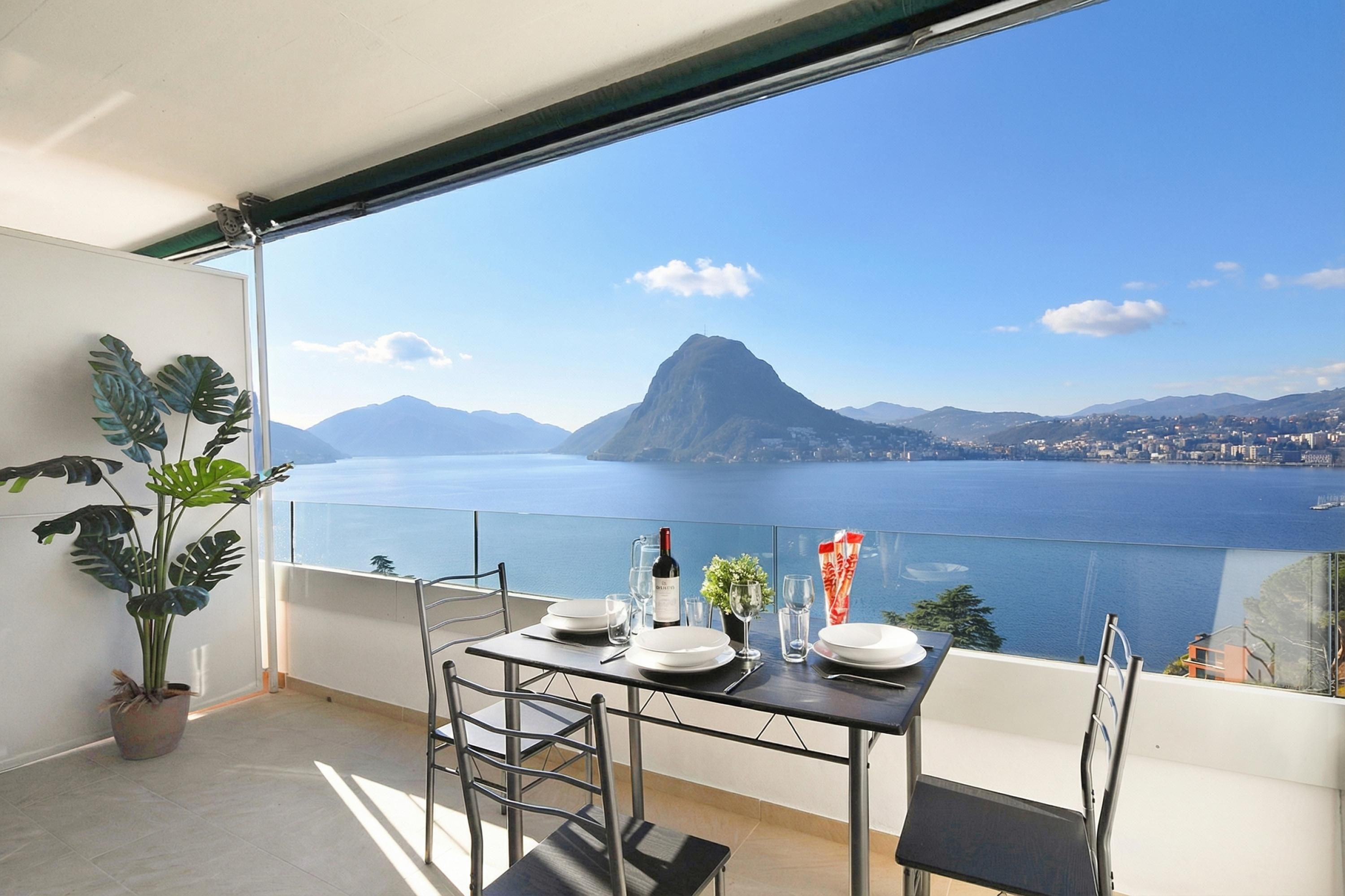 Un balcon avec une table dressée pour le dîner, un pot de fleurs et une vue sur une montagne et une étendue d’eau.