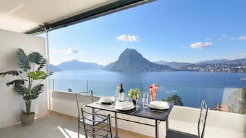 Un balcone con tavolo apparecchiato per mangiare, una pianta in vaso e vista su una montagna e un corpo d'acqua.