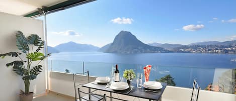 A balcony with a table set for dining, a potted plant, and a view of a mountain and body of water.