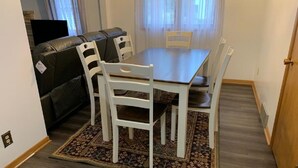 Ein Essbereich mit einem Holztisch und vier weißen Stühlen, einem gemusterten Teppich und einer dunkelbraunen Ledersofa im Hintergrund.