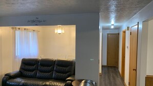 Ein Zimmer mit einer schwarzen Ledersofa, einem kleinen runden Tisch und einer wandmontierten Lampe. Rechts ist ein Korridor mit Holztüren zu sehen.
