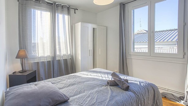 Une chambre à coucher comprenant un lit, une table de chevet, une lampe, une armoire et une fenêtre donnant sur des immeubles.