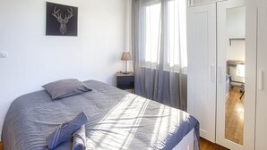 Une chambre à coucher avec un lit, une table de chevet, une lampe, une armoire et une fenêtre avec des rideaux.