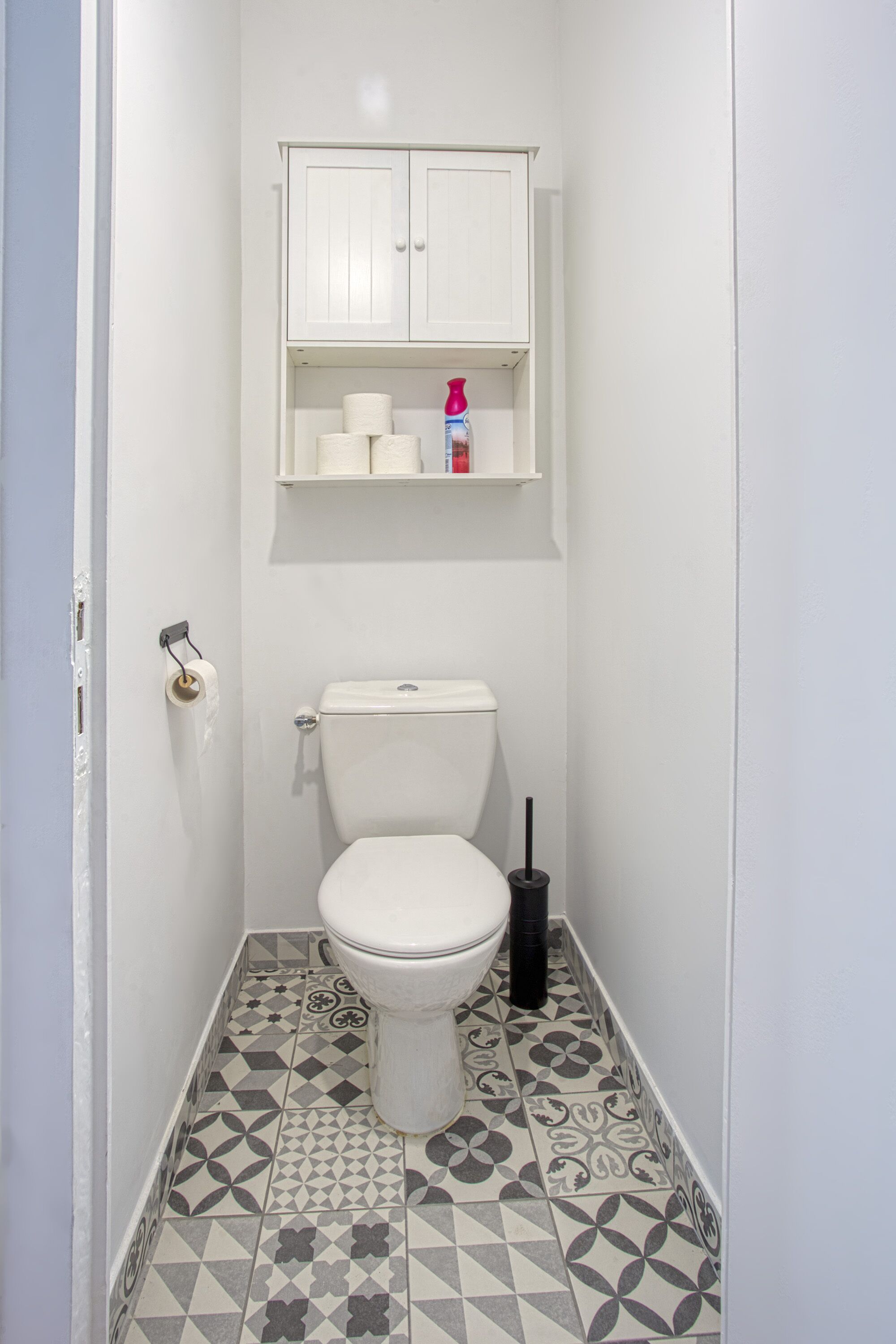 Une petite salle de bain avec des toilettes, des carreaux de sol à motifs, un meuble blanc au-dessus des toilettes et un rouleau de papier toilette accroché au mur.