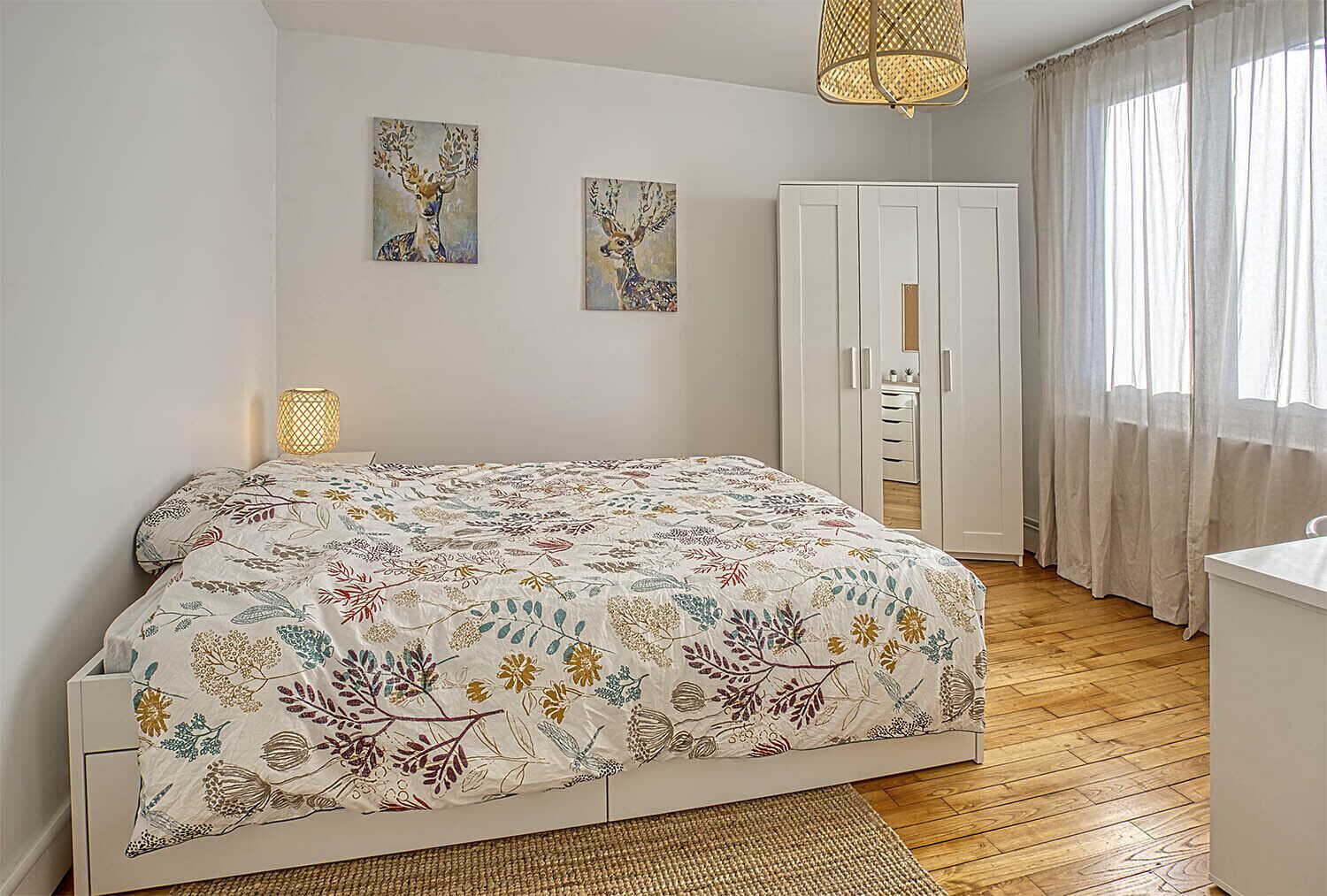 A bedroom with a floral-patterned bedspread, a wardrobe, a bedside lamp, and two wall-mounted paintings.