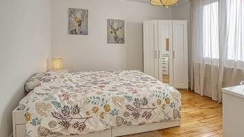 A bedroom with a floral-patterned bedspread, a wardrobe, a bedside lamp, and two wall-mounted paintings.