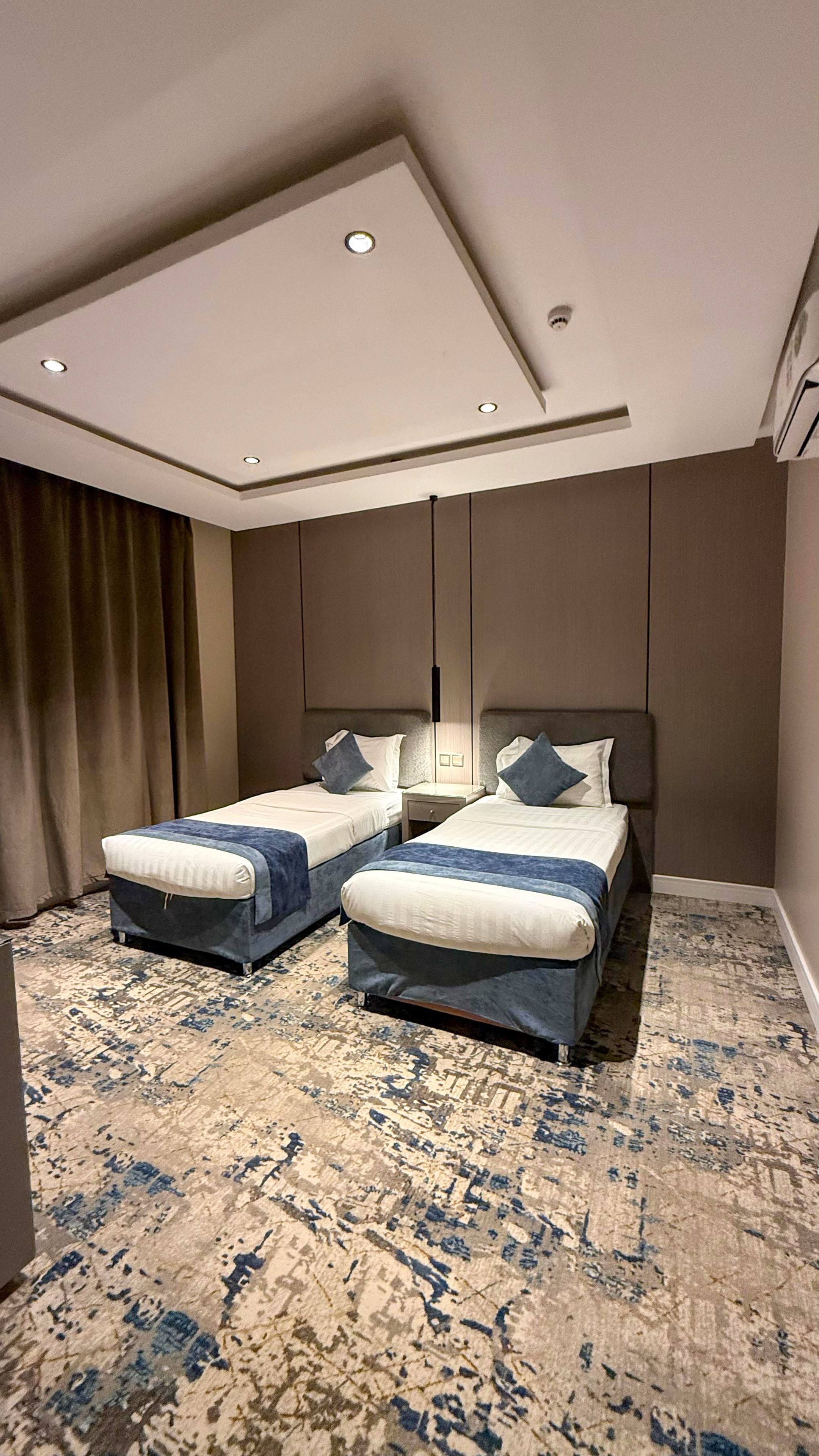 A hotel room with two beds, a patterned carpet, and a ceiling with recessed lighting.