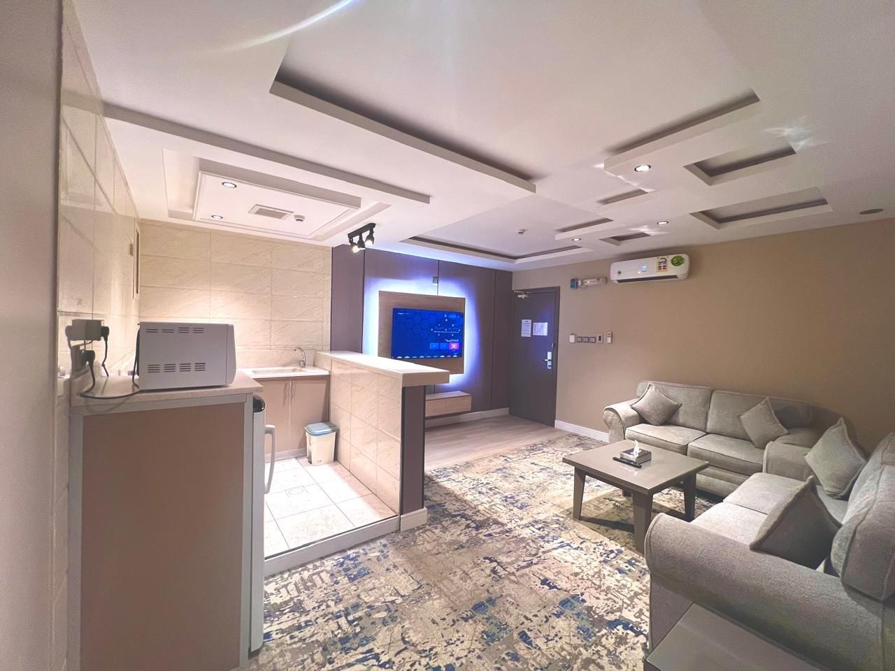 A modern living room with a flat-screen TV, a sofa, a coffee table, and a small kitchen area with a microwave.