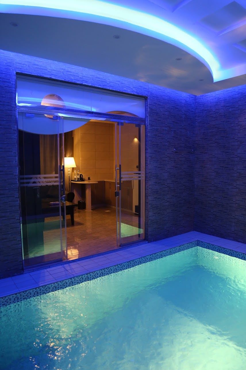 An indoor swimming pool with a glass wall leading to a room with a chair and a table.