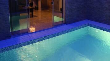 An indoor swimming pool with a glass wall leading to a room with a chair and a table.