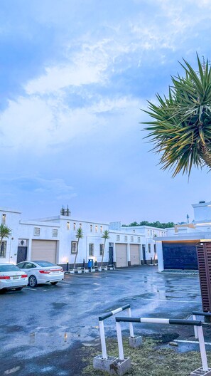 A parking lot with a white building in the background, a palm tree, and a car. - Manazel Aldana (Abha)