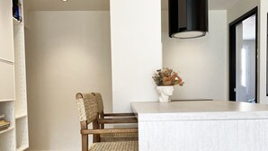 A modern kitchen with a white countertop, wicker chairs, a vase with dried flowers, and a black pendant light.