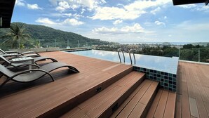 Une piscine à débordement avec une terrasse en bois et des chaises longues, offrant une vue sur le paysage urbain et les collines.