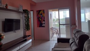 A living room with a flat-screen TV, a sofa, a potted plant, and a colorful painting on the wall. - Rua Maria Tognini 296 (Praia Grande)