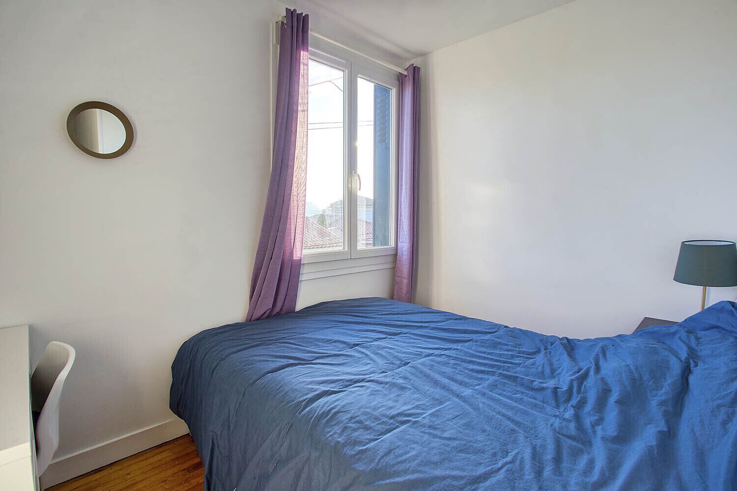 A bedroom with a bed, a window with purple curtains, a round mirror on the wall, and a lamp on the right.