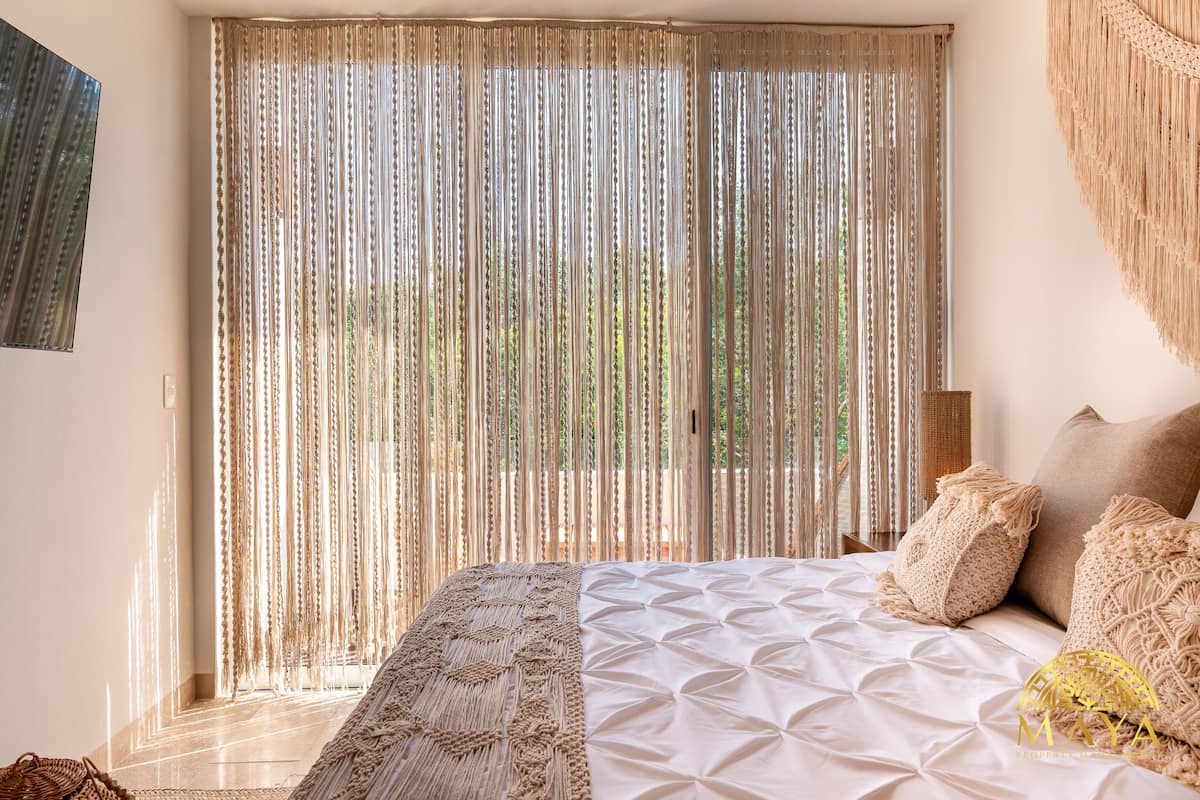 A bedroom with a large bed, beige curtains, and a woven throw blanket.