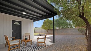 A covered patio area with two wooden chairs and a small table, a black door, and a gravel ground. - Newly Renovated Home, 1 mi Downtown - 3 BR 2 Bath (Chandler)