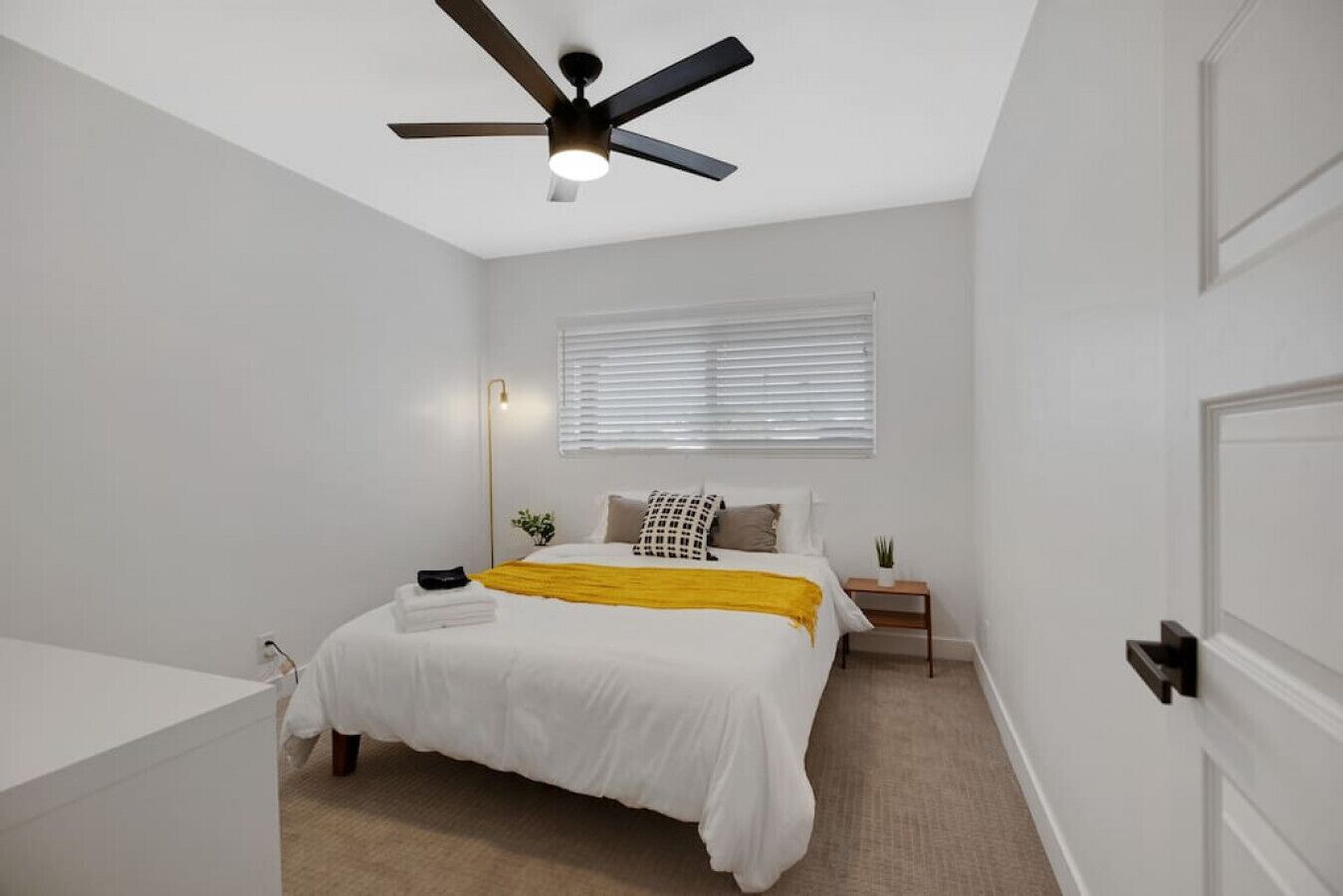 A bedroom with a bed, a bedside table, a nightstand, a ceiling fan, and a window with blinds.