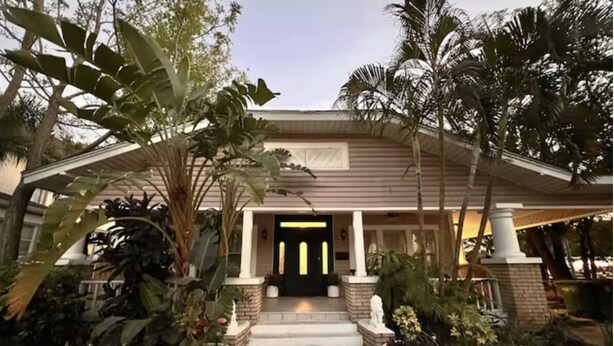 Une maison dotée d’un porche à l’avant, entourée de plantes tropicales et de palmiers.
