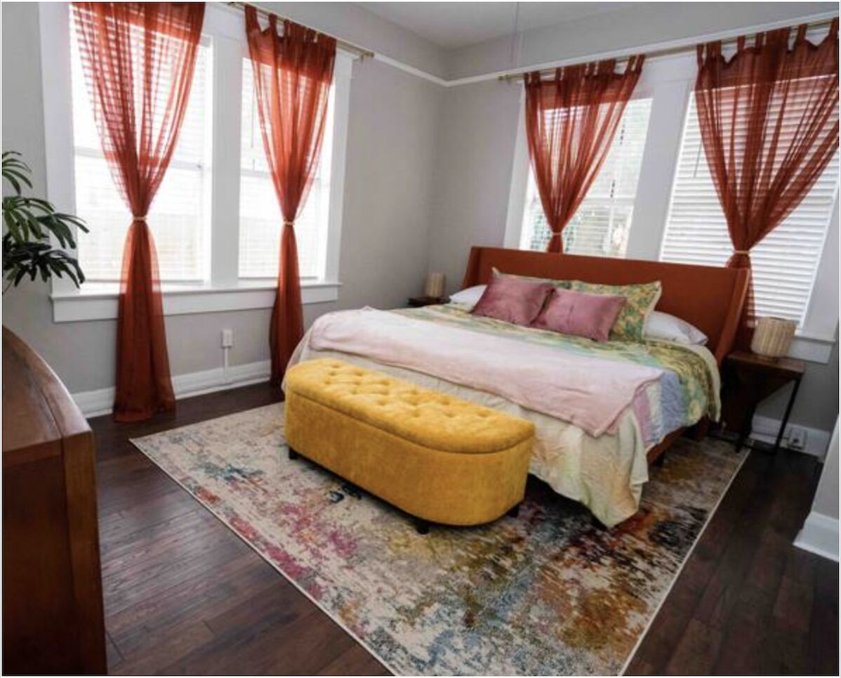 Un dormitorio con cama, un reposapiés amarillo, una alfombra estampada y cortinas rojas.