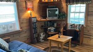 L’intérieur d’un chalet accueillant, avec un plancher en bois, un canapé bleu, une table en bois, un téléviseur, une bibliothèque et une fenêtre avec des stores.