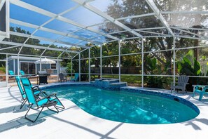 Une piscine couverte avec un toit en verre, entourée de chaises longues et d’un espace barbecue.
