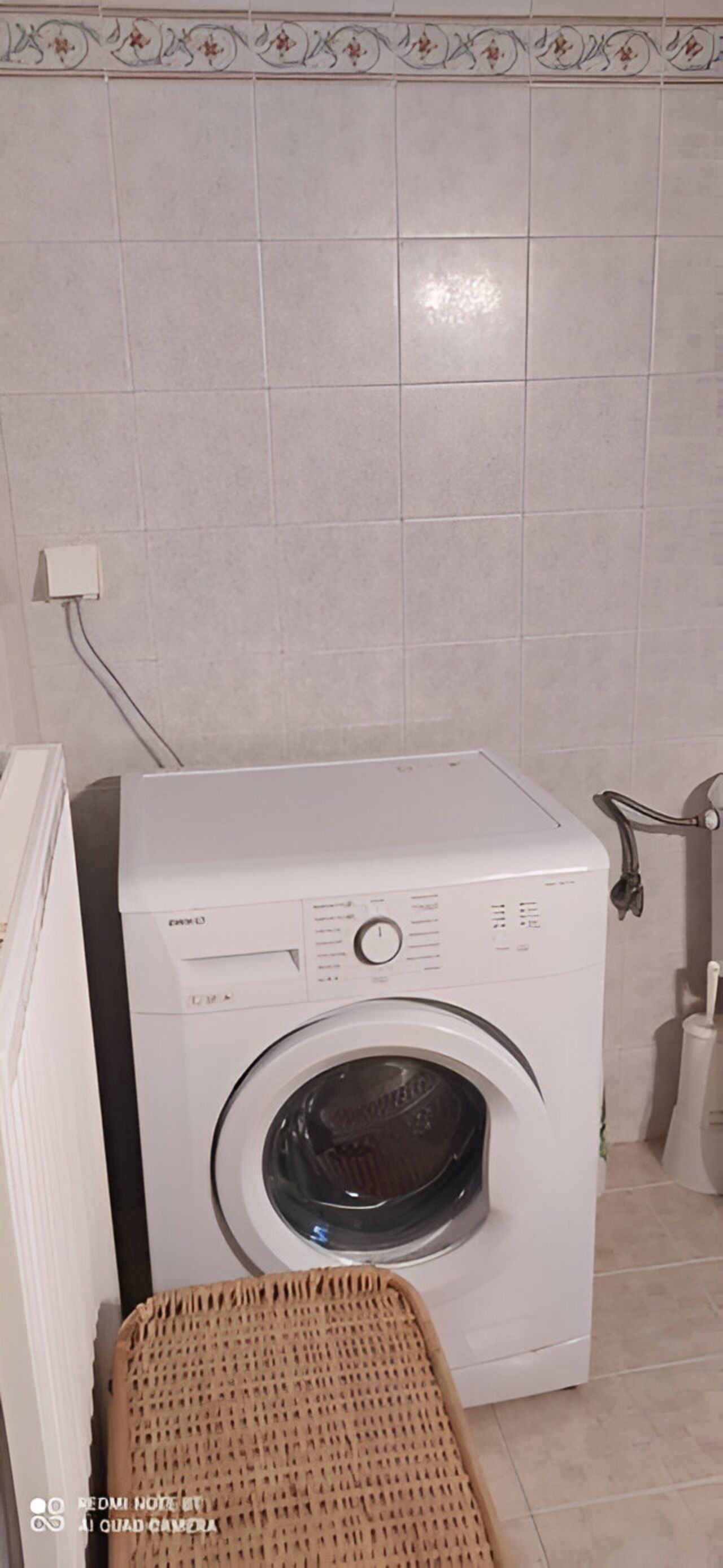 A front-loading washing machine in a laundry room with tiled walls and a wicker basket.