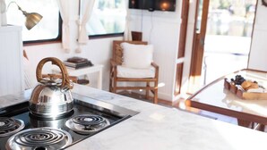 A kitchen with a gas stove, a kettle, and a marble countertop. There is a chair and a table with a basket of bread in the background.