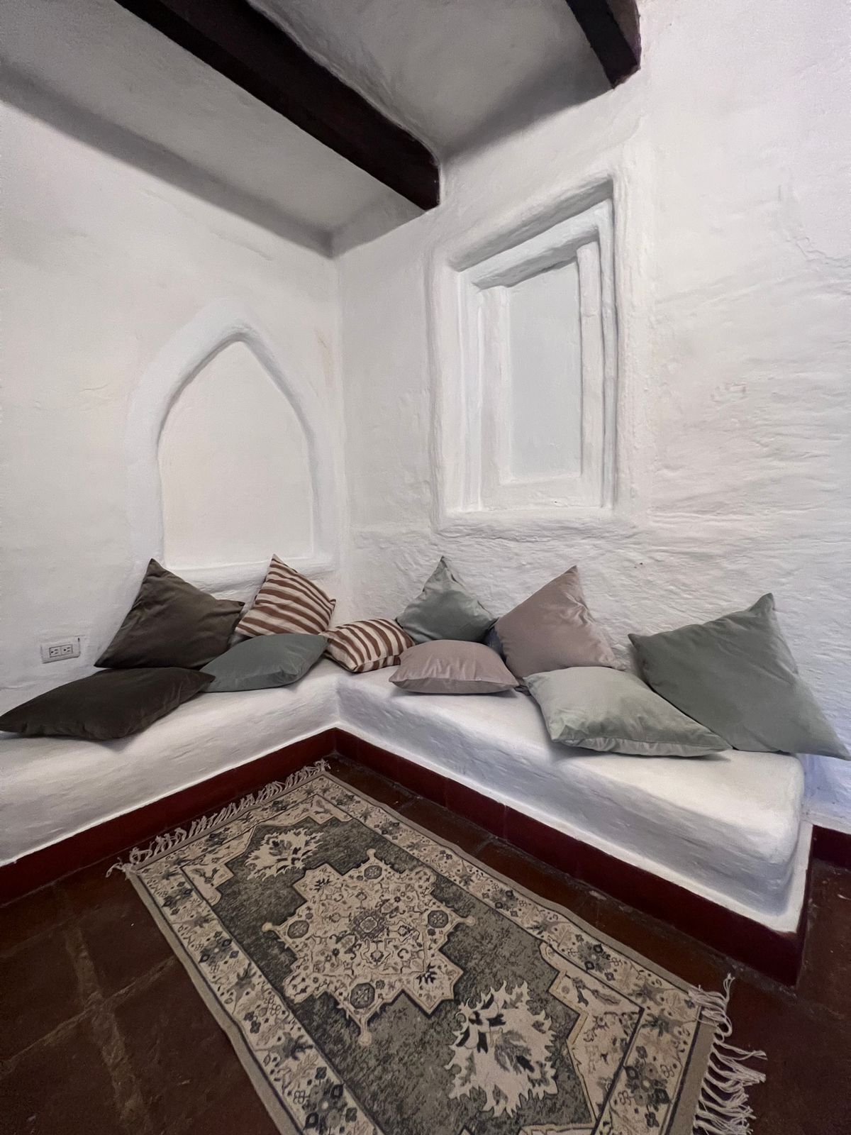 Una habitación con un banco blanco y bajo contra una pared blanca, decorada con varios cojines. Una alfombra estampada cubre el suelo.