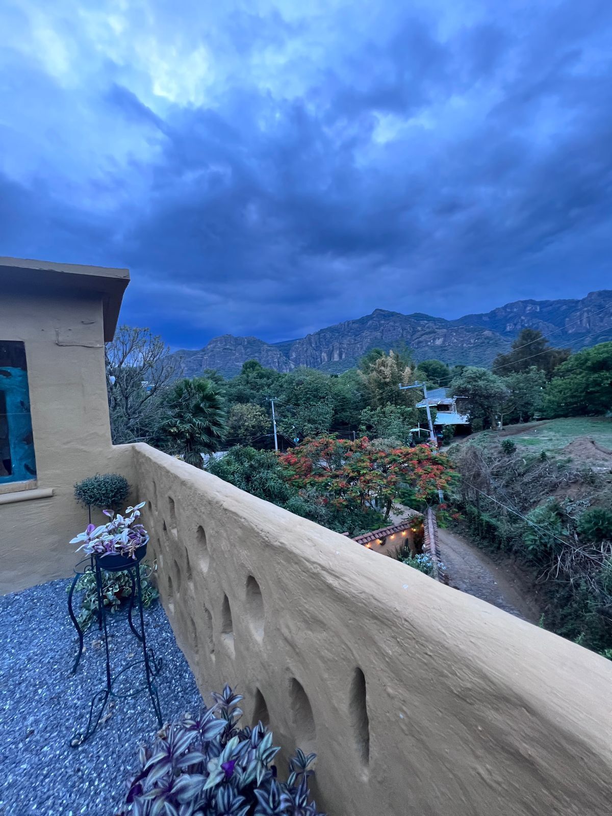 Un balcone con vista su un paesaggio montuoso, un edificio con facciata beige e un piccolo giardino con piante in vaso.