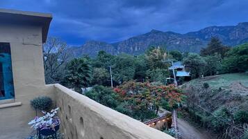 Un balcone con vista su un paesaggio montuoso, un edificio con facciata beige e un piccolo giardino con piante in vaso.
