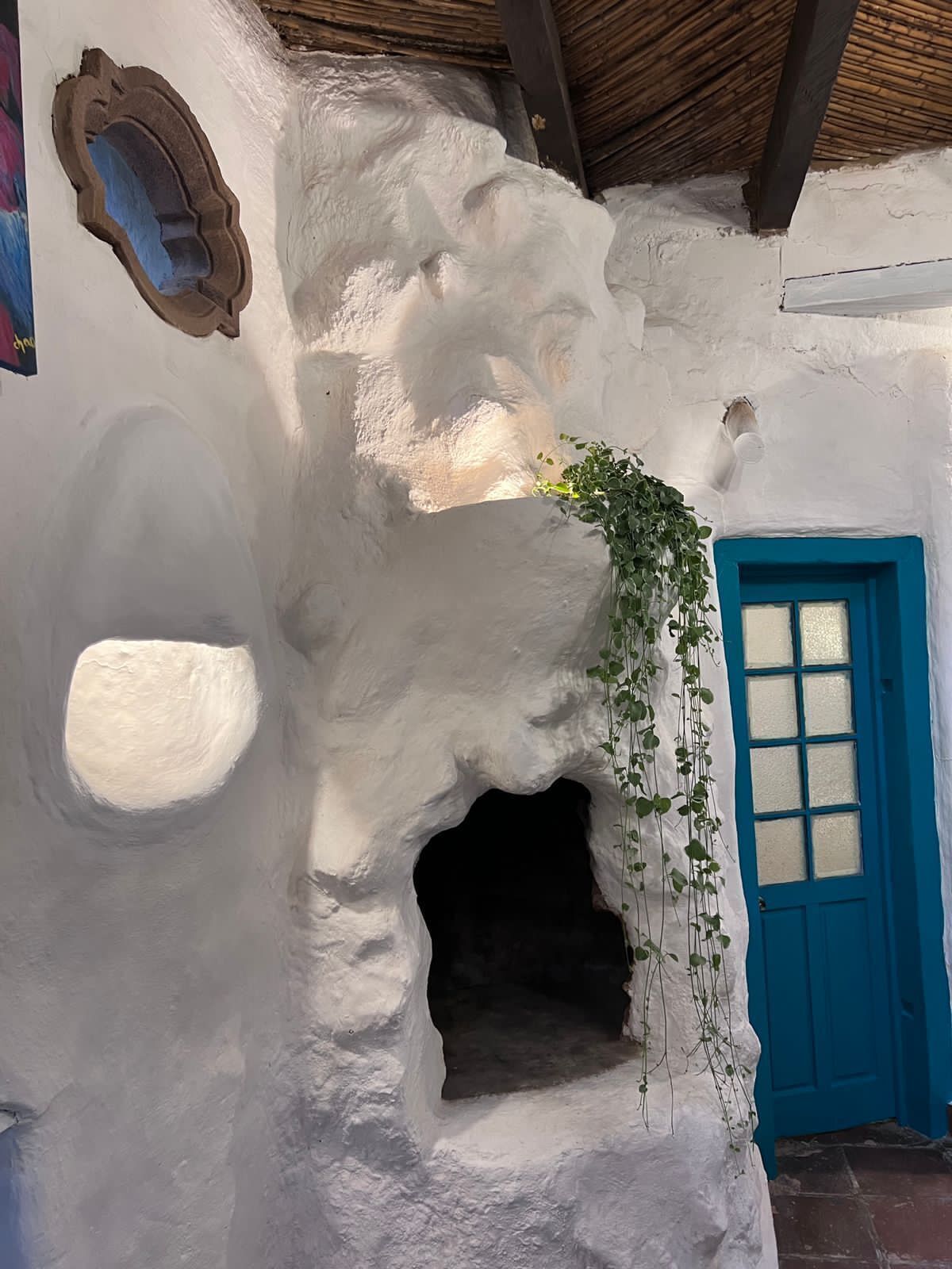 Un interior rústic amb una porta blava, una finestra petita i una planta penjant.