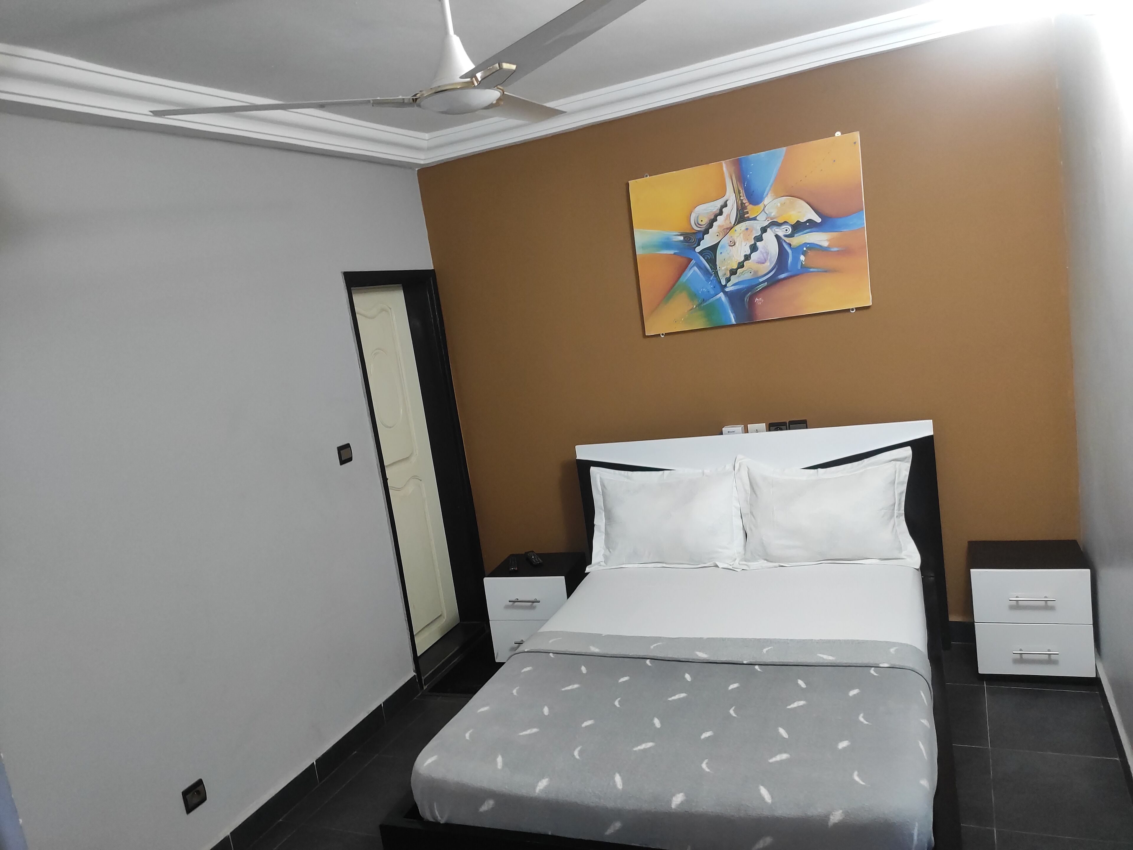 A modern hotel room with a bed, bedside tables, a ceiling fan, and a painting on the wall.