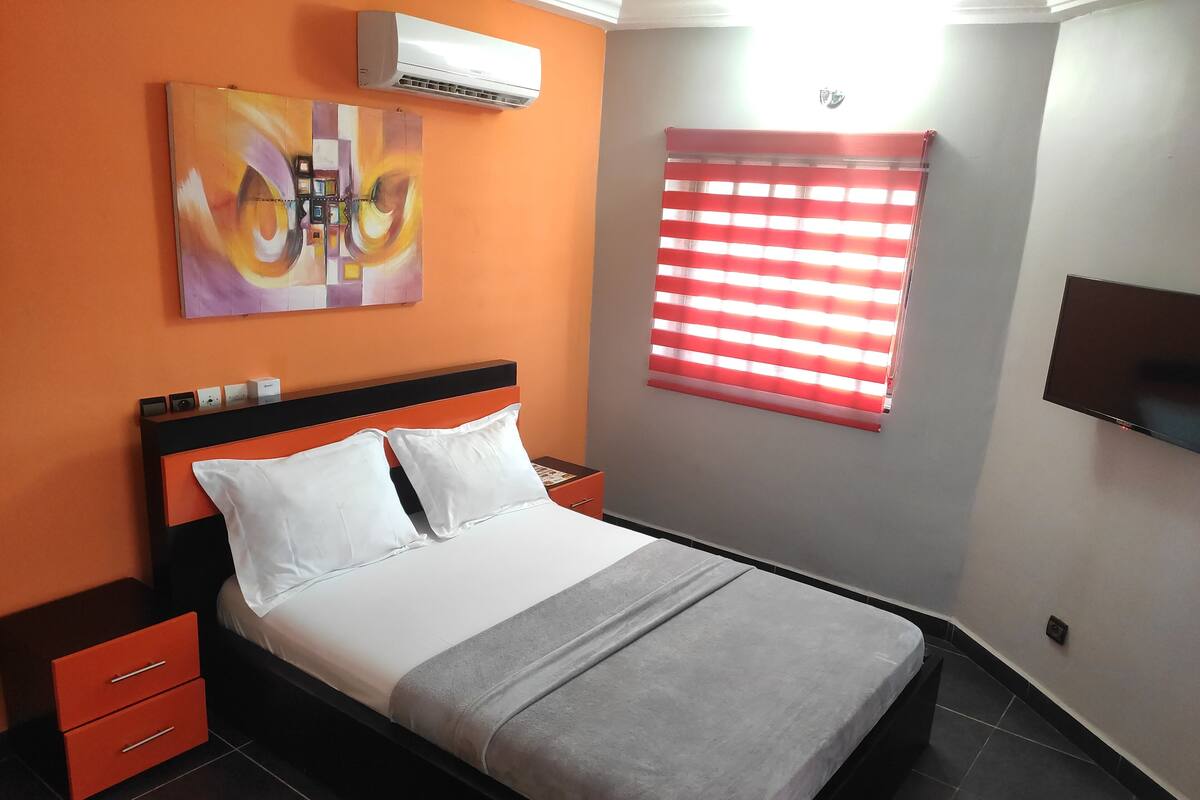 A neatly made bed with white and grey bedding, a red nightstand with drawers, a wall-mounted TV, and a window with red blinds.