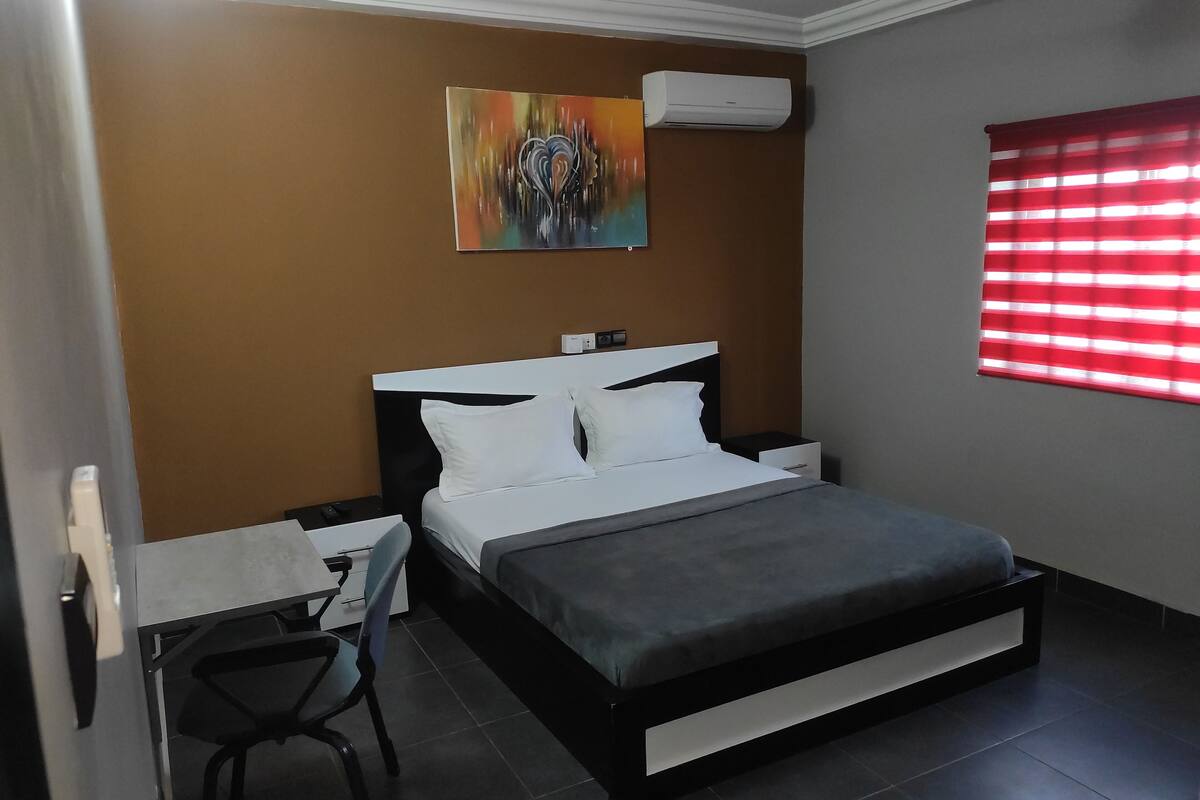 A bedroom with a bed, a desk, a chair, an air conditioner, and a window with blinds.