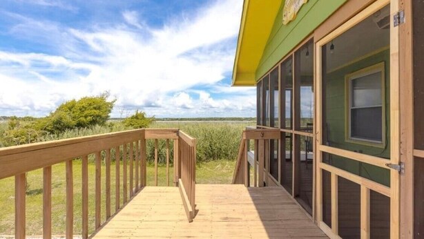 A wooden deck with a railing, a green building with large windows, and a view of a grassy field under a partly cloudy sky. - Overlook the Intracoastal and walk to the beach! (North Topsail Beach)