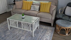 A living room with a beige sofa, a glass coffee table, a wicker chair, a patterned rug, and a painting on the wall. - Overlook the Intracoastal and walk to the beach! (North Topsail Beach)
