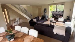 Un salon spacieux avec un canapé aux couleurs sombres, une table à manger en bois agrémentée de sets de table en fibres naturelles et un escalier avec une rampe noire.