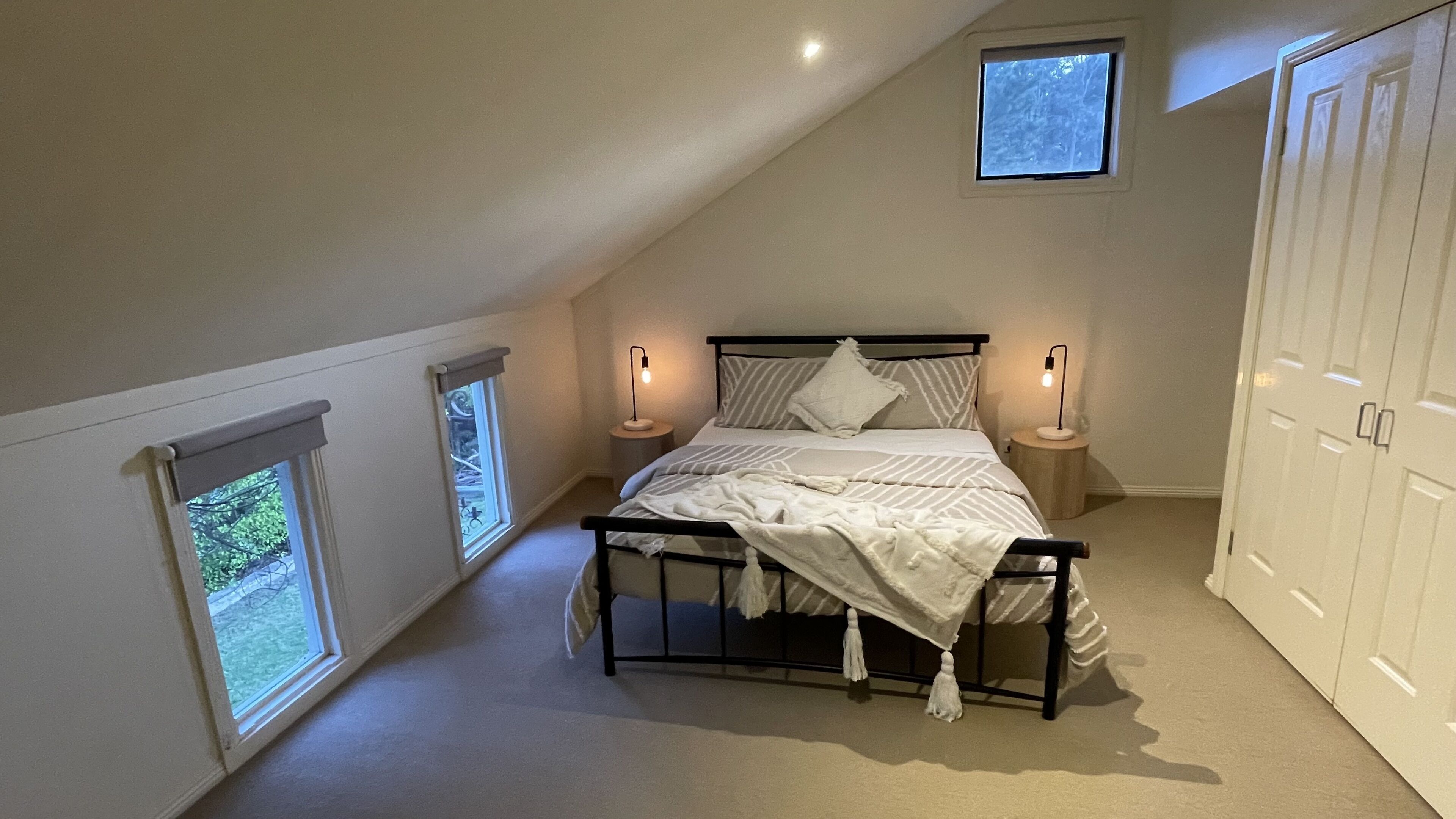 A bedroom with a sloped ceiling, a bed with a white comforter, two bedside lamps, and a wardrobe.