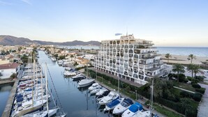 A marina with boats docked, a large white building with multiple balconies, and a beach in the background.