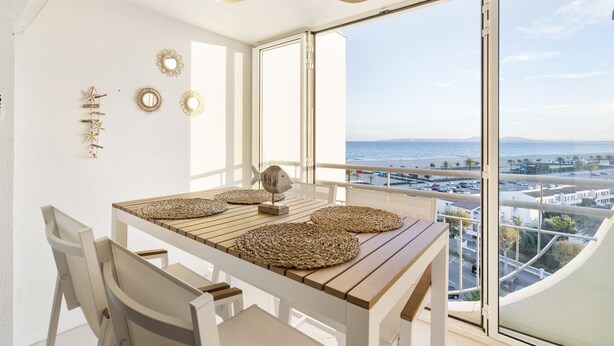 A balcony with a table and chairs, a ceiling fan, decorative wall mirrors, and a view of the ocean and cityscape.