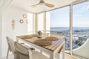 Un balcón con mesa y sillas, un ventilador de techo, espejos decorativos en la pared y vista al océano y al paisaje urbano.