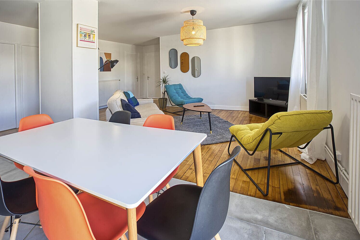 En moderne stue med spisebord, farverige stole, et fjernsyn og en hængende lampe.
