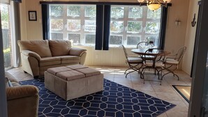 Una sala de estar acogedora con un sofá color beige, un taburete a juego y una mesa de comedor con sillas. Amplias ventanas con persianas permiten la entrada de luz natural.