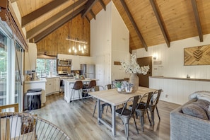 A spacious kitchen with wooden beams, a dining area with a table and chairs, and a large window overlooking a forested area. - Cozy & Chic Lake Arrowhead Cabin w/ Deck! (Lake Arrowhead)