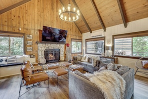 A spacious living room with a stone fireplace, leather seating, and large windows offering views of the outdoors. - Cozy & Chic Lake Arrowhead Cabin w/ Deck! (Lake Arrowhead)
