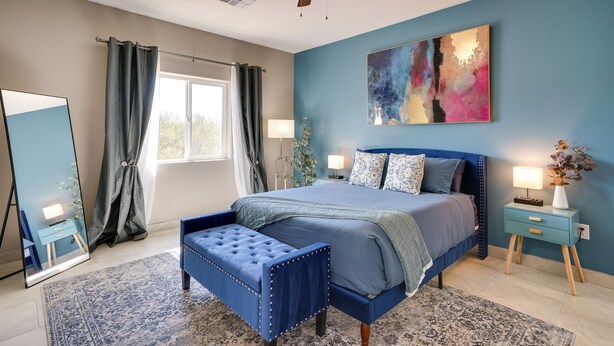 A bedroom with a bed, a ceiling fan, a large mirror, a nightstand with a lamp, and a window with curtains. - Tucson Vacation Rental w/ Community Pool! (Tucson)