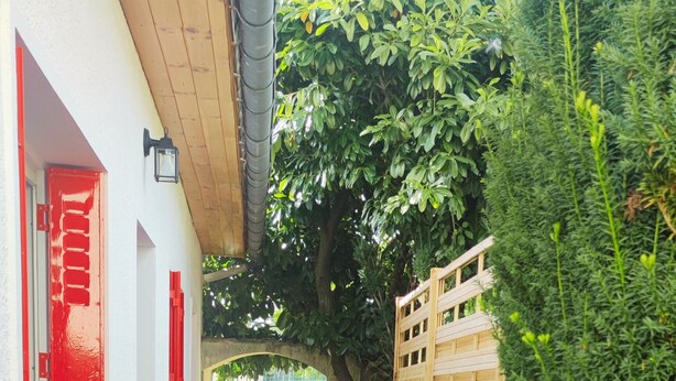 Ein schmaler Betonweg, der von einem weißen Gebäude mit roten Fensterläden und einem Holzzaun gesäumt wird.