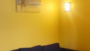 Ein Zimmer mit einem dunkelgrauen Sofa, einer Deckenleuchte, einem Wandbild und einer kleinen beleuchteten Wandpaneele.